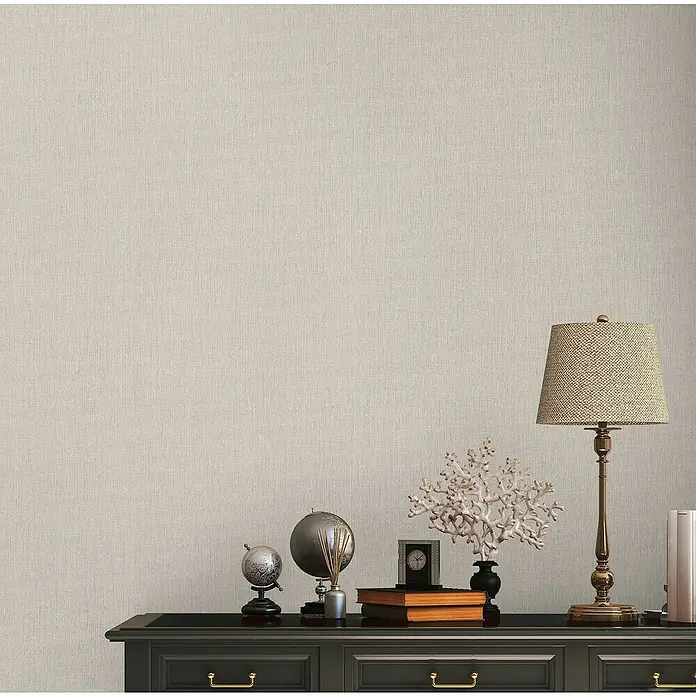 A.S. Création Stories of Life Vliestapete - Graubeige, Uni, 10,05 x 0,53 m - Dekorative Tischleuchte mit Lampenschirm und Zierobjekten auf einer schwarzen Kommode, die eine warme und elegante Atmosphäre schafft.