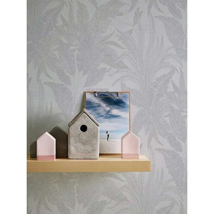 A.S. Création Stories of Life Vliestapete Dschungel - Grau-Creme-Gold, Floral, 10,05 x 0,53 m - Dekoratives Regal mit Zement- und rosa Häusern, einer Fotoklemme mit Strandmotiv vor einer Tapete mit Blattmuster.