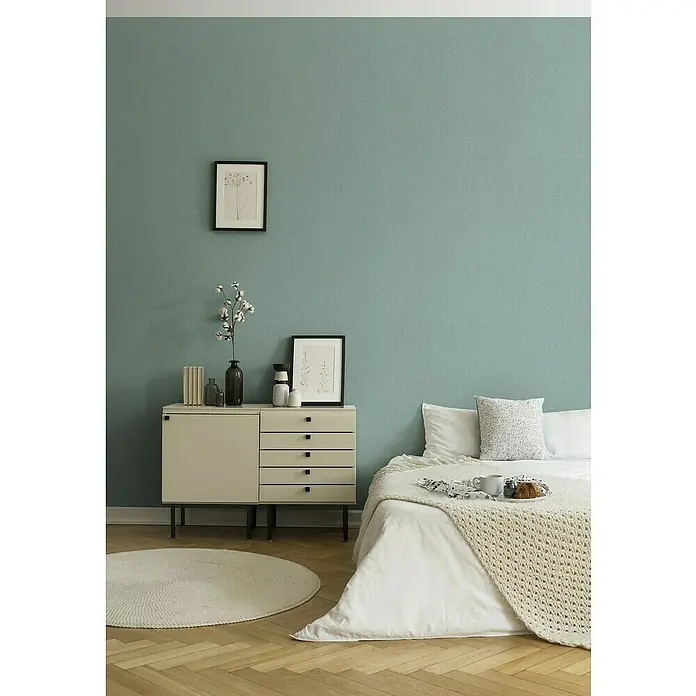 A.S. Création Stories of Life Vliestapete - Salbeigrün, Uni, 10,05 x 0,53 m - Schlafzimmer mit einem Bett mit weißer Bettwäsche, einer Kommode und einem runden Teppich. Das Zimmer strahlt eine ruhige und gemütliche Atmosphäre aus.
