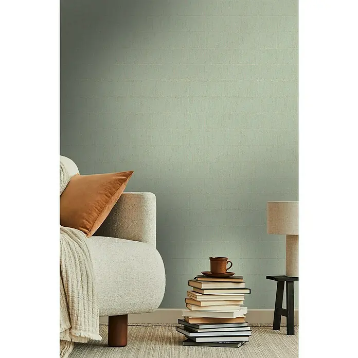 A.S. Création Jade 2 Vliestapete - Goldgrün, Grafisch, 10,05 x 0,53 m - Nahaufnahme eines Wohnzimmers mit grün gestreifter Wand, Sessel, Kissen, Decke, Lampe und Bücherstapel mit Tasse. Die ruhige Atmosphäre lädt zum Verweilen ein.