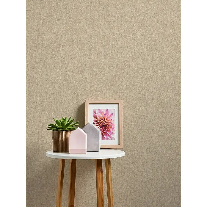 A.S. Création Famous Garden Vliestapete - Beige, Uni, 10,05 x 0,53 m - Nahaufnahme eines dekorativen Beistelltisches mit Zimmerpflanze, Hausdeko und einem Bilderrahmen mit einer rosa Blume vor einer beigen Wand.