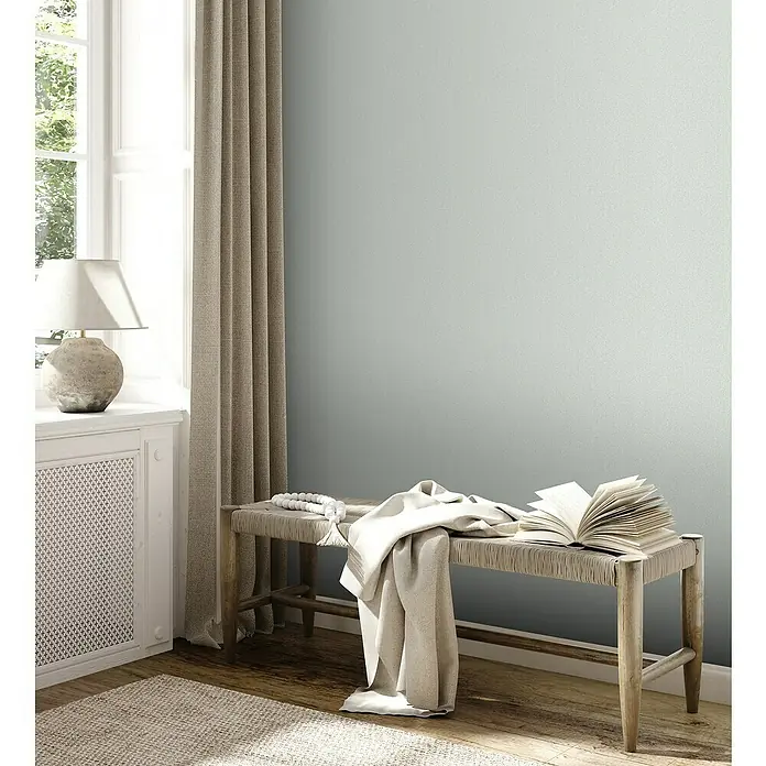 A.S. Création Jade 2 Vliestapete - Mintgrün, Uni, 10,05 x 0,53 m - Eine Bank mit einem Buch und einer Decke in einem gemütlichen, hellen Raum mit neutraler Farbpalette, die eine friedliche Atmosphäre schafft.