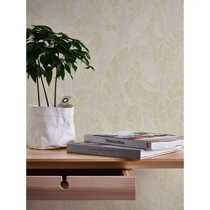 A.S. Création Jade 2 Vliestapete Blätter - Weiß-Beige-Gold, Floral, 10,05 x 0,53 m - Nahaufnahme eines Tisches mit einer Pflanze, Büchern und einer Schublade vor einer Tapete mit Blattmuster.
