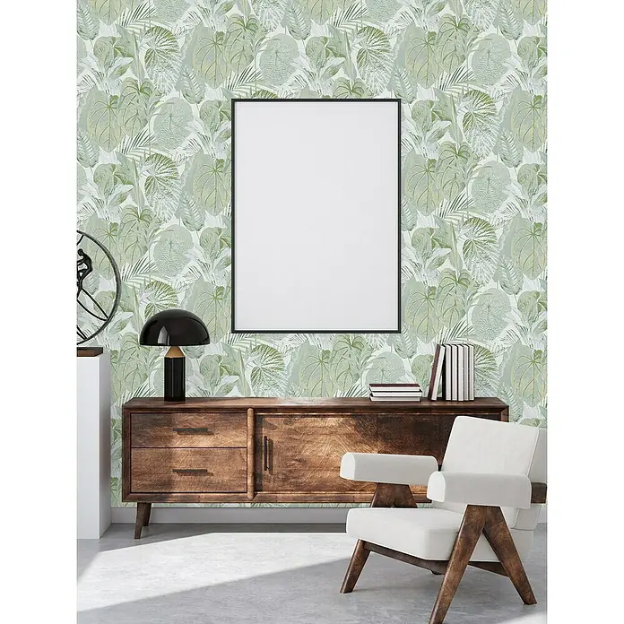 A.S. Création Famous Garden Vliestapete Dschungel - Grün-Weiß-Grau, Floral, 10,05 x 0,53 m - Wohnzimmer mit Holzkommode, Lampe, Büchern, Sessel und einem gerahmten, leeren Poster an der Wand mit Blättern.