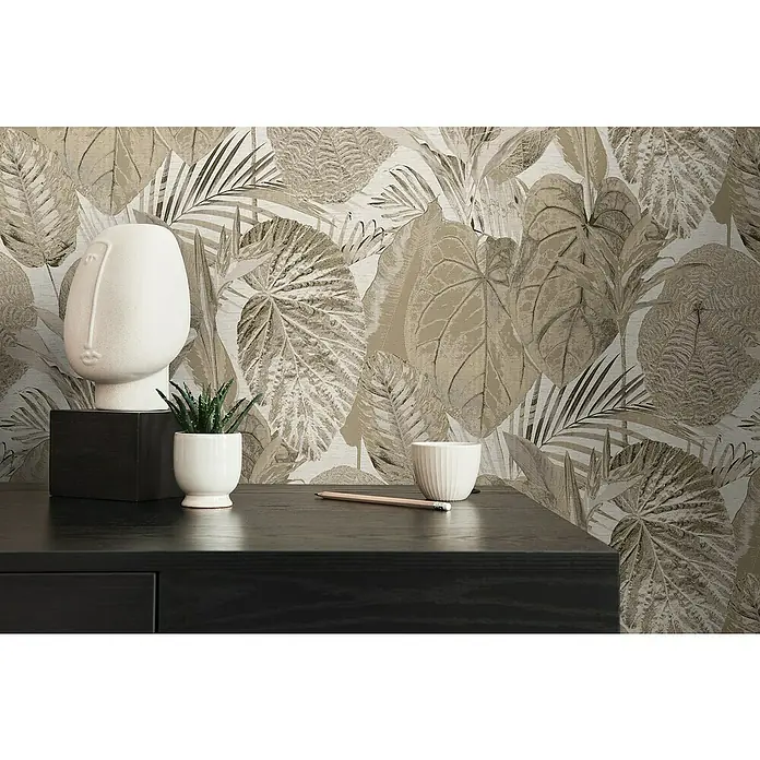 A.S. Création Famous Garden Vliestapete Dschungel - Beige-Taupe, Floral, 10,05 x 0,53 m - Nahaufnahme von Dekoartikeln, darunter eine Skulptur, eine Topfpflanze und ein Stift auf einem dunklen Tisch vor einer gemusterten Wand.