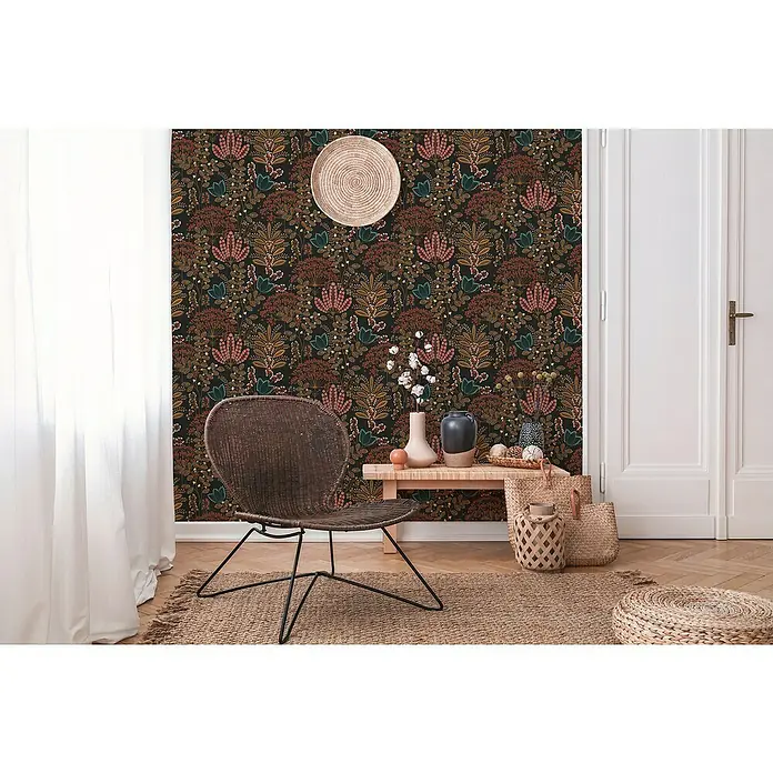 A.S. Création Famous Garden Vliestapete - Schwarz-Bunt-Petrol, Floral, 10,05 x 0,53 m - Gemütliches Interieur mit einem Rattansessel auf einem gemusterten Teppich vor einer floralen Tapete.