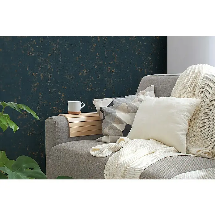 A.S. Création Jade 2 Vliestapete - Dunkelblau-Gold, Uni, 10,05 x 0,53 m - Nahaufnahme eines grauen Sofas mit weißen Kissen und einer hellen Decke, ergänzt durch einen praktischen Armlehnentisch mit einer Tasse.