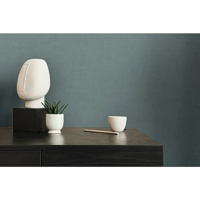 A.S. Création Jade 2 Vliestapete - Blau-Grün, Uni, 10,05 x 0,53 m - Nahaufnahme eines Schreibtisches mit einer minimalistischen Skulptur, einer kleinen Pflanze, einer Tasse und einem Bleistift vor einer strukturierten Wand.