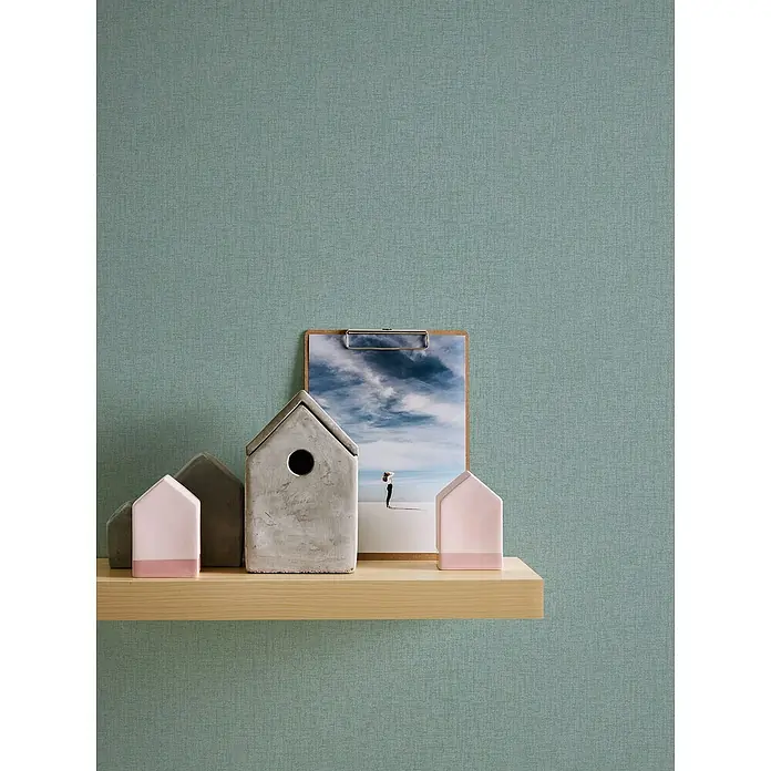 A.S. Création Stories of Life Vliestapete - Salbeigrün, Uni, 10,05 x 0,53 m - Ein Regal mit Dekorationsgegenständen wie Häusern in Betonoptik und einem Bild mit einem bewölkten Himmel.