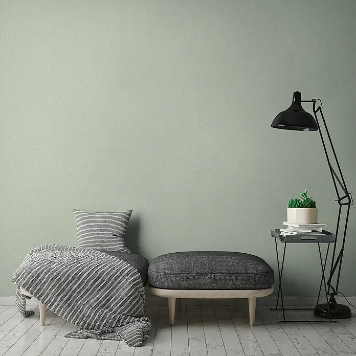 A.S. Création Jade 2 Vliestapete - Pastellgrün, Uni, 10,05 x 0,53 m - Einladender Raum mit grau gestreiftem Kissen und Decke auf einer Bank, ergänzt durch einen eleganten Hocker und eine Stehlampe mit Pflanze auf einem Beistelltisch.