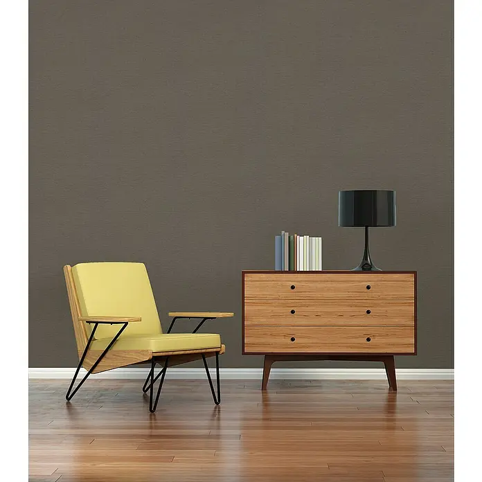 A.S. Création Famous Garden Vliestapete - Braun, Uni, 10,05 x 0,53 m - Das Bild zeigt einen gelben Sessel und eine Holzkommode mit einer Lampe und Büchern darauf vor einer braunen Wand.