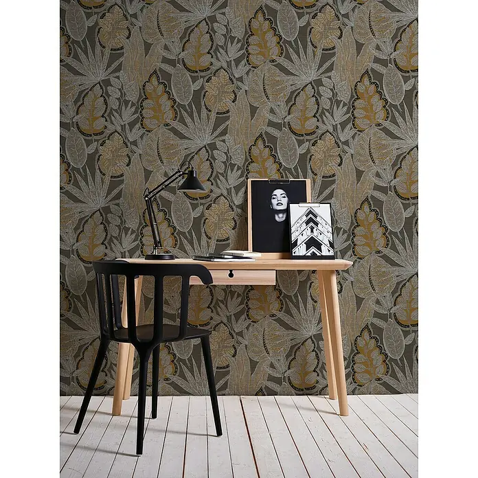 A.S. Création Famous Garden Vliestapete Blatt - Braun-Orange-Weiß, Floral, 10,05 x 0,53 m - Nahaufnahme eines Schreibtischs mit dekorativer Tischleuchte, Holzrahmen und einem schwarzen Stuhl vor einem blattgemusterten Hintergrund.