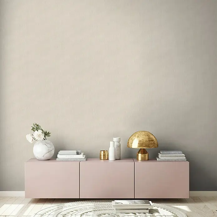 A.S. Création Stories of Life Vliestapete - Hellbeige, Uni, 10,05 x 0,53 m - Nahaufnahme eines stilvollen Interieurs mit einem rosa Sideboard, einer goldenen Lampe und dekorativen Elementen vor einer neutralen Wand.
