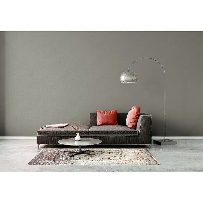 A.S. Création Famous Garden Vliestapete - Grau, Uni, 10,05 x 0,53 m - Das graue Sofa mit den roten Kissen, dem Tisch und der Lampe sorgt für eine gemütliche Atmosphäre im Raum. Der Teppich rundet das Gesamtbild ab.