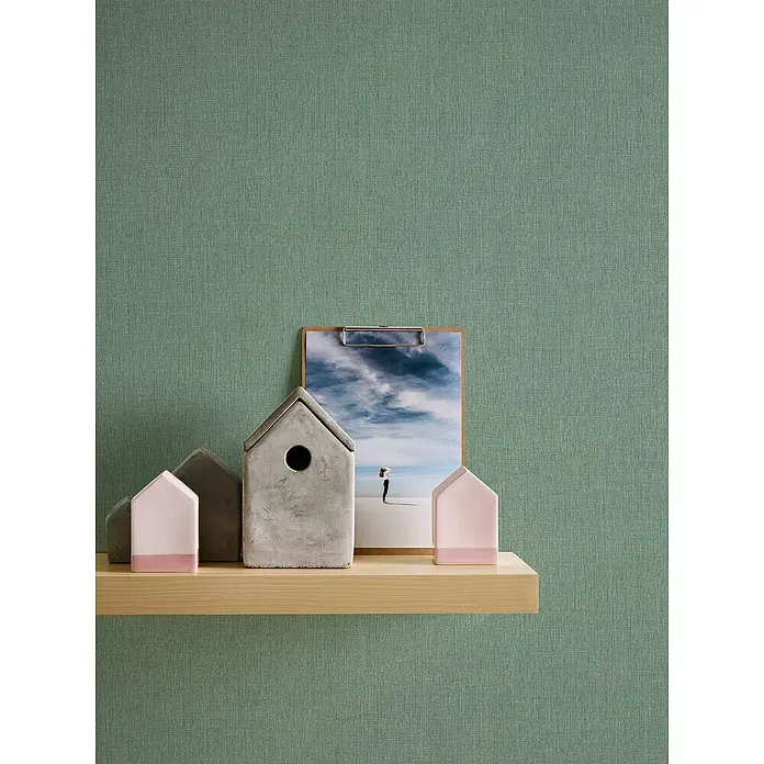 A.S. Création Stories of Life Vliestapete - Dunkelgrün, Uni, 10,05 x 0,53 m - Nahaufnahme von dekorativen Miniaturhäusern aus Beton und einem gerahmten Bild auf einem Holzregal vor einem grünen Hintergrund.