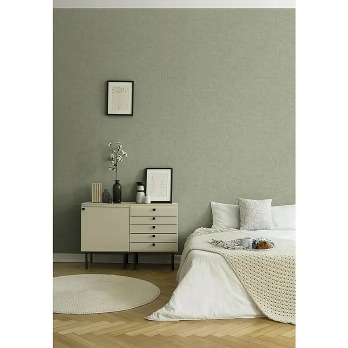 A.S. Création Jade 2 Vliestapete - Grün, Uni, 10,05 x 0,53 m - Das Schlafzimmer verfügt über eine moderne Kommode mit Schubladen, eine stilvolle Lampe und ein gerahmtes Kunstwerk an einer olivgrünen Wand.