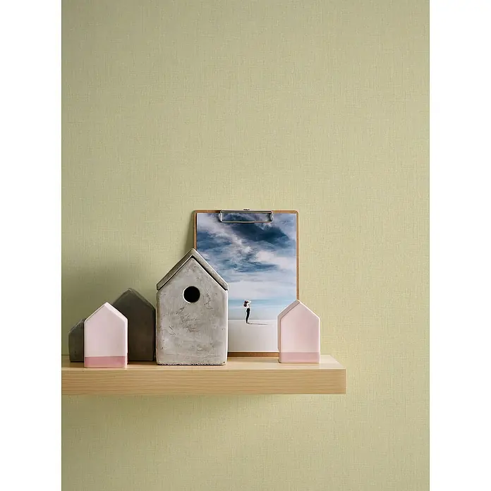 A.S. Création Stories of Life Vliestapete - Gelb-Grün, Uni, 10,05 x 0,53 m - Dekorative Elemente auf einem Regal: Zementhäuser, rosa Akzente und eine Fotografie auf einer Holzplatte sorgen für eine gemütliche Atmosphäre.