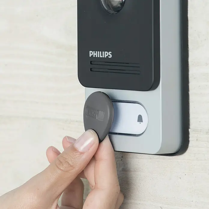 Philips WelcomeEye RFID-Badge - 2er Set, Passend für: Philips Welcome Eye System - Nahaufnahme einer Hand, die einen grauen RFID-Tag an einen intelligenten Philips Türspion hält, um das Gerät zu aktivieren.