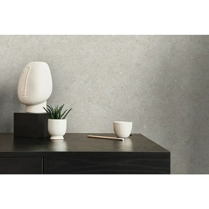 A.S. Création Stories of Life Vliestapete Used-Look - Beige-Silber, Betonoptik, 10,05 x 0,53 m - Nahaufnahme einer schwarzen Kommode mit einer weißen Skulptur, einer Pflanze und einer Tasse vor einer grauen Wand.