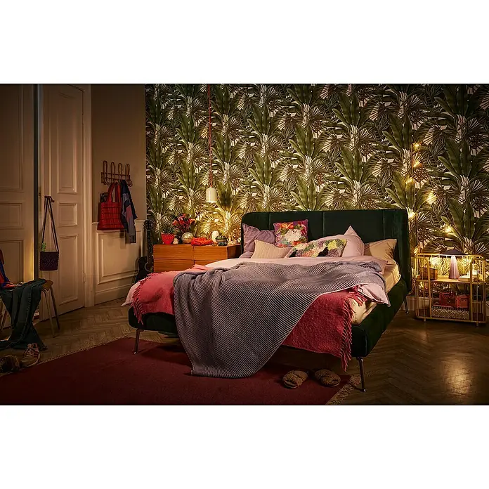 A.S. Création Stories of Life Vliestapete - Grün-Cremeweiß-Beige, Floral, 10,05 x 0,53 m - Schlafzimmer mit einem grünen Bett mit Decken und Kissen, einem Holztisch und tropischer Tapete.