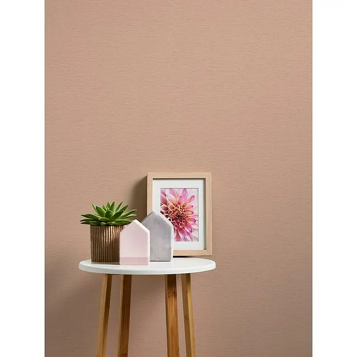 A.S. Création Famous Garden Vliestapete - Rosa, Uni, 10,05 x 0,53 m - Nahaufnahme eines kleinen Beistelltisches mit Dekorationen: einer Pflanze, Deko-Häusern und einem Bilderrahmen mit einem Blumenmotiv.