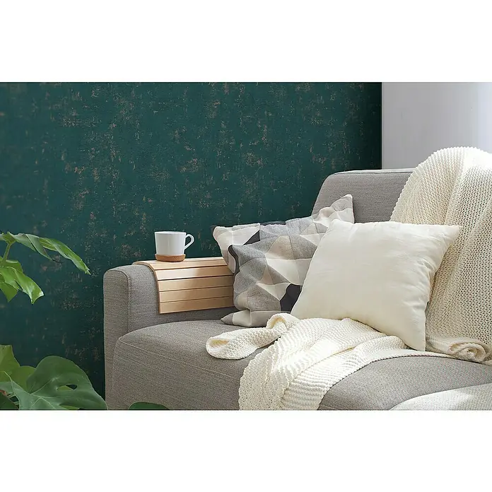 A.S. Création Jade 2 Vliestapete - Petrol-Gold, Uni, 10,05 x 0,53 m - Das Bild zeigt ein graues Sofa mit Kissen, Decke und einem Holztablett auf der Armlehne, auf dem eine weiße Tasse steht. Zimmerpflanze sichtbar.