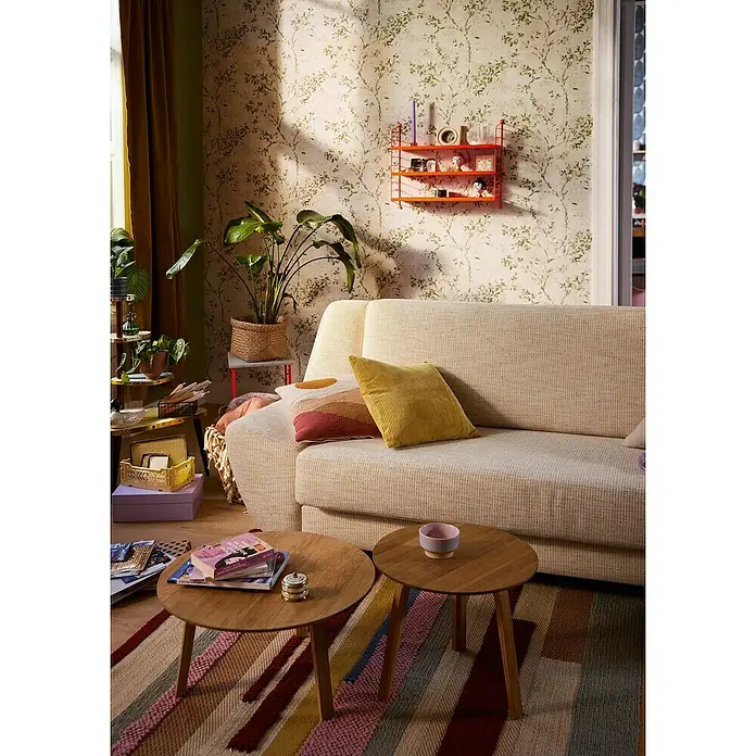 A.S. Création Stories of Life Vliestapete Zweig - Beige-Grün-Gold, Floral, 10,05 x 0,53 m - Gemütliches Wohnzimmer mit beigem Sofa, Holztischen, farbenfrohem Teppich und Pflanzendeko. Die orangefarbene Wandregal bietet zusätzlichen Stauraum.