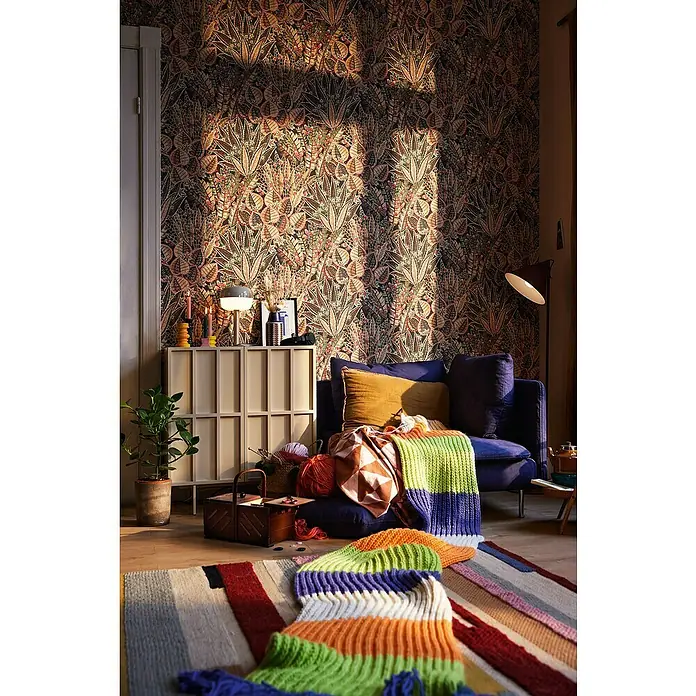 A.S. Création Stories of Life Vliestapete Blumen - Braun-Rot-Schwarz, Floral, 10,05 x 0,53 m - Gemütliches Interieur mit einem blauen Sessel, einem bunten Teppich und einem Sideboard mit Strickutensilien vor einer gemusterten Tapete.