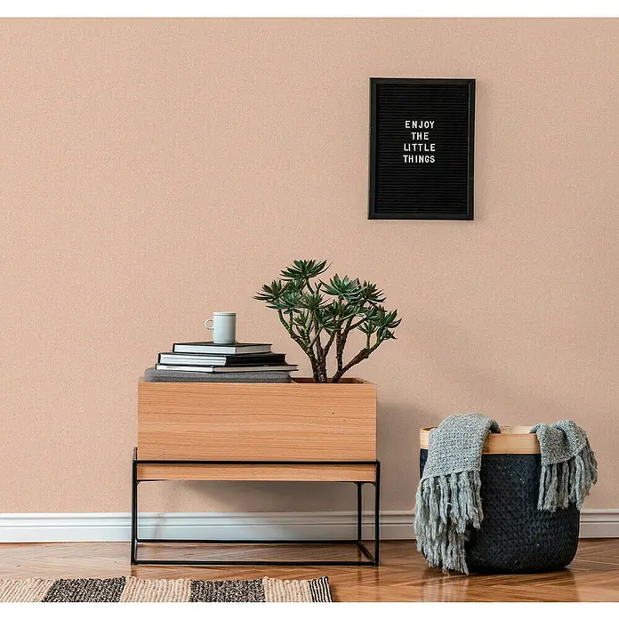 A.S. Création Famous Garden Vliestapete - Orange-Rosa, Uni, 10,05 x 0,53 m - Nahaufnahme einer Zimmerpflanze auf einem Holztisch mit Büchern, einer Tasse, einem Korb mit einer Decke und einer dekorativen Tafel an der Wand.