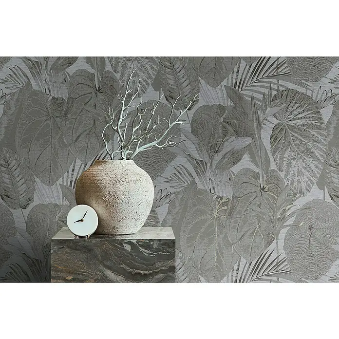 A.S. Création Famous Garden Vliestapete Dschungel - Grau-Anthrazit, Floral, 10,05 x 0,53 m - Produktfoto mit einer Vase und einer Uhr auf einem Marmorpodest, vor einem Hintergrund mit tropischen Pflanzen.