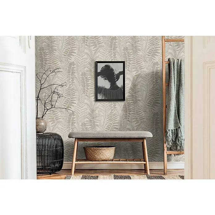 A.S. Création Famous Garden Vliestapete - Grau-Beige-Taupe, Floral, 10,05 x 0,53 m - Elegante Bank mit grauem Kissen, Weidenkorb und dunklem Beistelltisch auf einem gestreiften Teppich vor einer Wand mit Blattmuster.