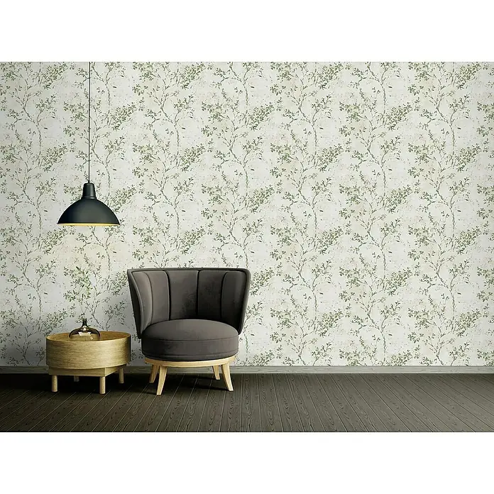 A.S. Création Stories of Life Vliestapete Zweig - Beige-Grün-Gold, Floral, 10,05 x 0,53 m - Ein grauer Sessel und ein runder Beistelltisch mit goldener Dekoration, beleuchtet von einer Hängelampe vor einer Wand mit Blattmuster.