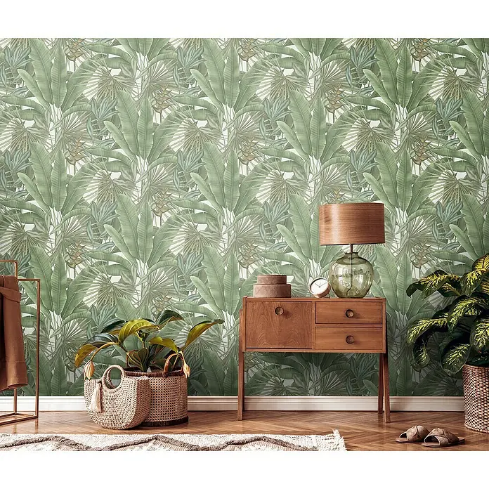 A.S. Création Stories of Life Vliestapete - Grün-Cremeweiß-Beige, Floral, 10,05 x 0,53 m - Detailaufnahme eines Raumes mit einem hölzernen Beistelltisch, einer Lampe und Pflanzen vor einer grünen Blättertapete.