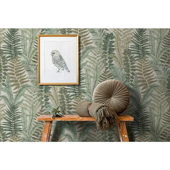 A.S. Création Famous Garden Vliestapete - Beige-Grün-Braun, Floral, 10,05 x 0,53 m - Dekorative Wand mit Farnmuster, verziert mit einem gerahmten Vogelbild, einem runden Kissen und einer Strickdecke auf einem Holztisch.
