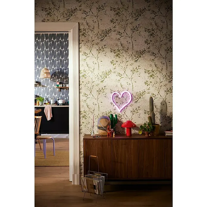 A.S. Création Stories of Life Vliestapete Zweig - Beige-Grün-Gold, Floral, 10,05 x 0,53 m - Wohnzimmer mit einem Sideboard aus Holz, auf dem ein Neonherz und verschiedene Dekoartikel stehen, Tapete mit Blätterdekor.