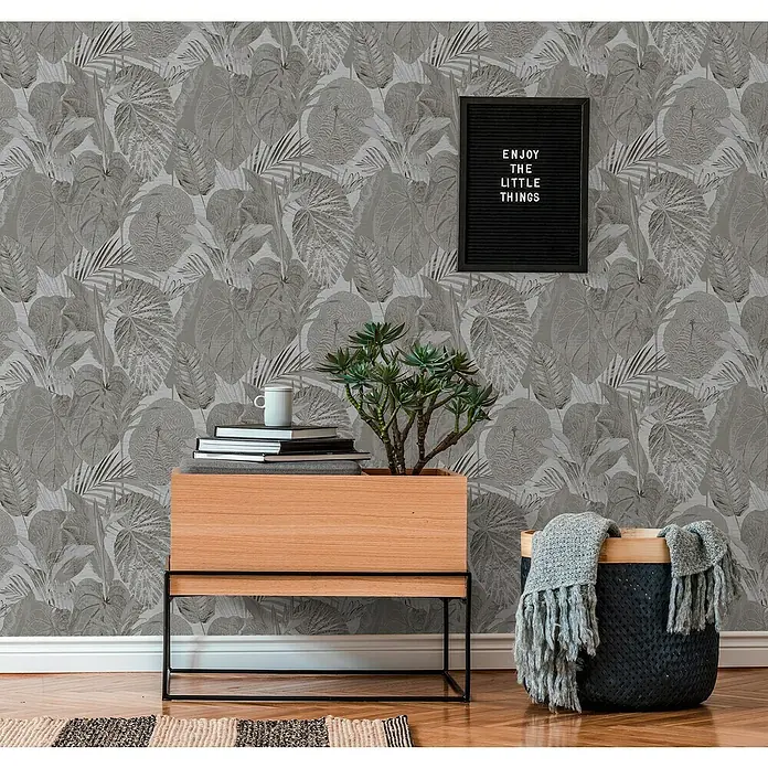 A.S. Création Famous Garden Vliestapete Dschungel - Grau-Anthrazit, Floral, 10,05 x 0,53 m - Ein schönes Interieur mit einem modernen Holzschrank, Bücherstapel, einer Grünpflanze, einem Korb mit einer Decke und einer Wandtafel.