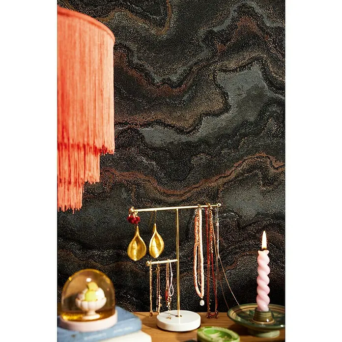 A.S. Création Stories of Life Vliestapete Marmoroptik Metallic-Effekt - Schwarz-Gold, Steinoptik, 10,05 x 0,53 m - Nahaufnahme von dekorativen Wohnaccessoires, darunter ein Schmuckständer, eine Schneekugel und eine rosa Kerze, vor einem gemusterten Hintergrund.