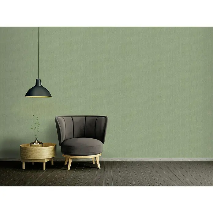 A.S. Création Stories of Life Vliestapete - Olivgrün, Uni, 10,05 x 0,53 m - Das Bild zeigt einen Sessel mit einem Beistelltisch und einer Lampe vor einer grünen Wand. Der Sessel ist grau und hat eine abgerundete Form.