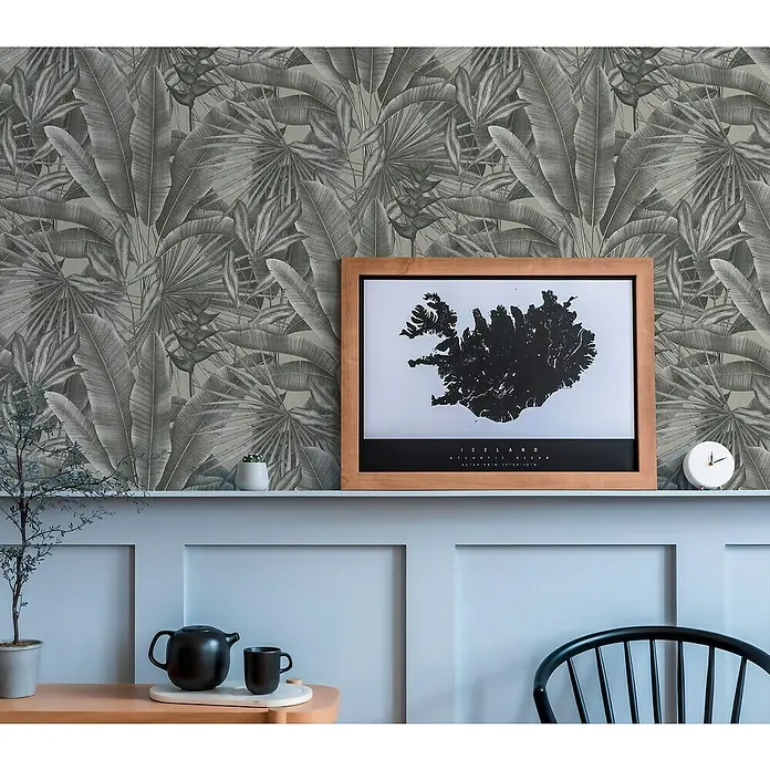 A.S. Création Stories of Life Vliestapete - Grau-Beige-Schwarz, Floral, 10,05 x 0,53 m - Zimmerdekor mit einer Karte von Island in einem Holzrahmen vor einer botanischen Tapete. Teekanne und Tasse stehen auf einem Beistelltisch.