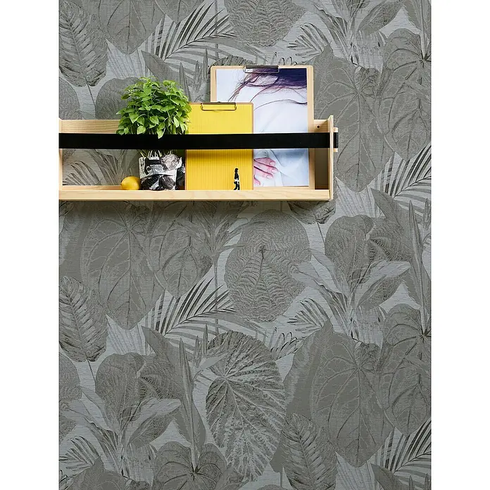 A.S. Création Famous Garden Vliestapete Dschungel - Grau-Anthrazit, Floral, 10,05 x 0,53 m - Das Bild zeigt ein hölzernes Wandregal mit einer Pflanze, einem gelben Notizbuch und Bildern, die auf einer Tapete mit tropischen Blättern platziert sind.