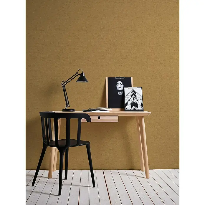 A.S. Création Famous Garden Vliestapete - Goldbraun-Gelb, Uni, 10,05 x 0,53 m - Ein minimalistischer Arbeitsplatz mit einem Schreibtisch und einem schwarzen Stuhl, beleuchtet durch eine Schreibtischlampe.