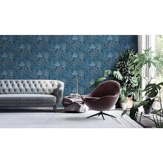 A.S. Création Famous Garden Vliestapete - Blau-Weiß-Grau, Floral, 10,05 x 0,53 m - Elegantes Wohnzimmer mit einem grauen Sofa, einem braunen Sessel und einer Wand mit floralem Muster. Zimmerpflanzen sorgen für eine lebendige Atmosphäre.