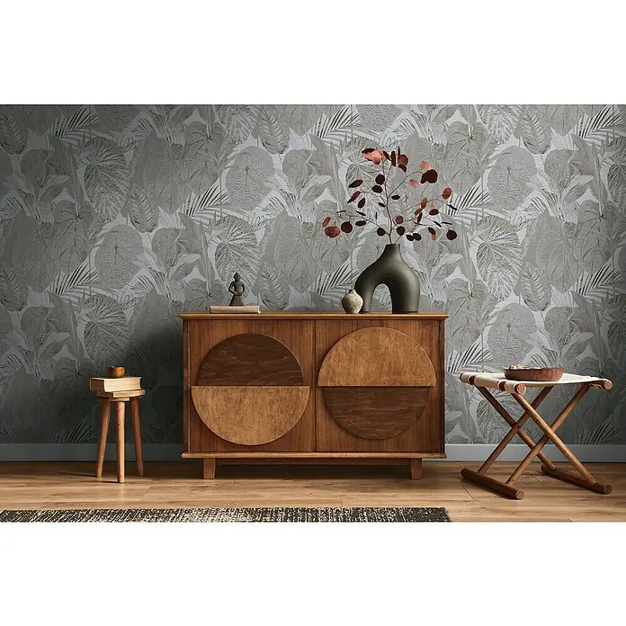 A.S. Création Famous Garden Vliestapete Dschungel - Grau-Anthrazit, Floral, 10,05 x 0,53 m - Zimmer mit hölzernem Sideboard mit runden Türen, Hocker und Faltstuhl vor einer grauen Blättertapete.