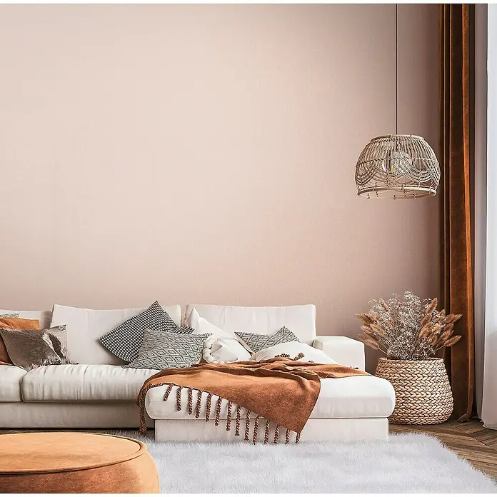 A.S. Création Famous Garden Vliestapete - Orange-Rosa, Uni, 10,05 x 0,53 m - Gemütliches Wohnzimmer mit weißem Sofa, Dekokissen und einer braunen Tagesdecke. Eine Rattanlampe hängt über dem Arrangement.