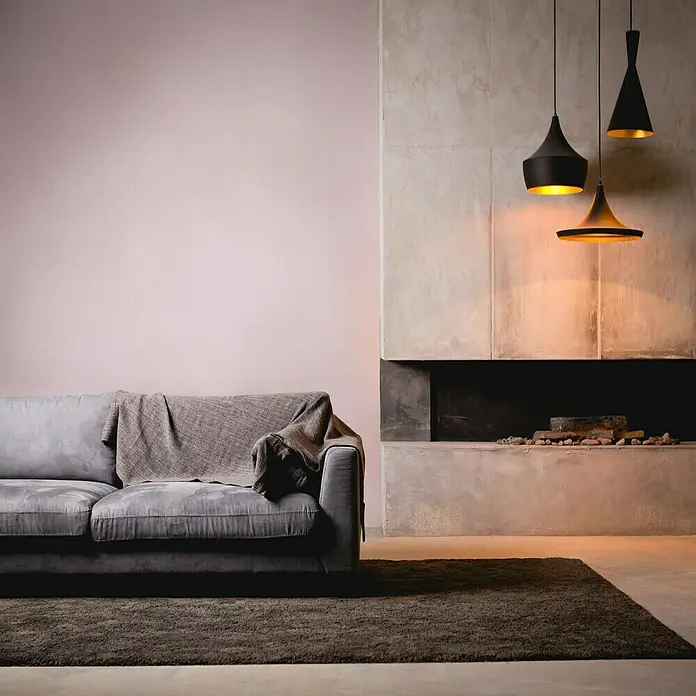 A.S. Création Stories of Life Vliestapete - Flieder, Uni, 10,05 x 0,53 m - Wohnzimmer mit modernem Sofa, dunklem Teppich, Kamin und drei stilvollen, schwarzen Pendelleuchten für eine gemütliche Atmosphäre.