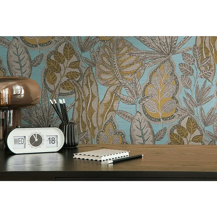 A.S. Création Famous Garden Vliestapete Blatt - Hellblau-Gelb-Braun, Floral, 10,05 x 0,53 m - Detailansicht eines Schreibtisches mit einer retro-analogen Uhr, einem Notizbuch und Stiften vor einer Wand mit tropischem Blattmuster.