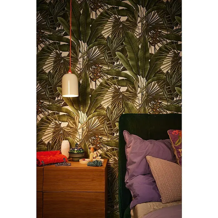 A.S. Création Stories of Life Vliestapete - Grün-Cremeweiß-Beige, Floral, 10,05 x 0,53 m - Nachttischlampe mit rotem Kabel vor einer tropischen Tapete, neben einem mit Kissen drapierten Bett.