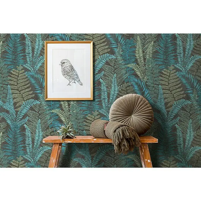 A.S. Création Famous Garden Vliestapete - Schwarz-Blau-Grün, Floral, 10,05 x 0,53 m - Ein Wanddekor mit Farnmuster, ergänzt durch einen goldenen Rahmen mit Vogelzeichnung und dekorativen Kissen auf einem kleinen Tisch.