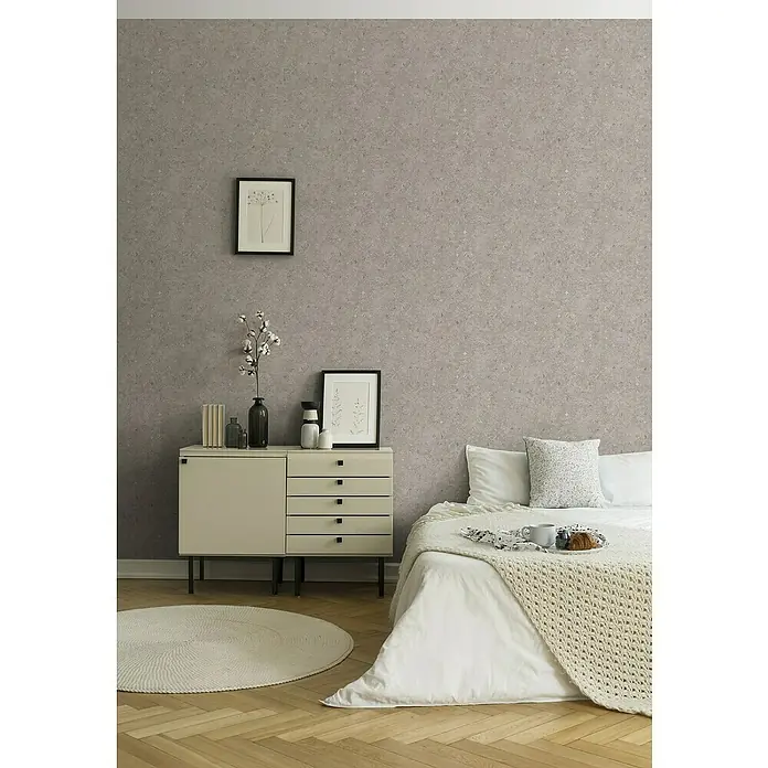 A.S. Création Stories of Life Vliestapete Used-Look - Beige-Braun, Betonoptik, 10,05 x 0,53 m - Schlafzimmer mit Kommode, Bett und rundem Teppich. Die Kommode in der Ecke bietet Stauraum, während das Bett zum Entspannen einlädt.