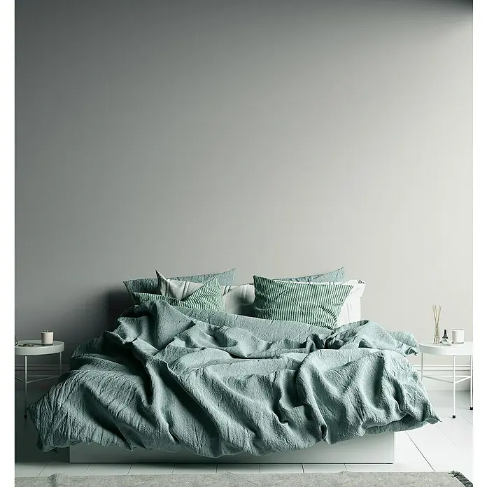 A.S. Création Famous Garden Vliestapete - Creme-Weiß, Uni, 10,05 x 0,53 m - Gemütliches Schlafzimmer mit einem großen Bett, das mit einer lässigen, hellgrünen Leinenbettwäsche und gestreiften Kissen dekoriert ist.